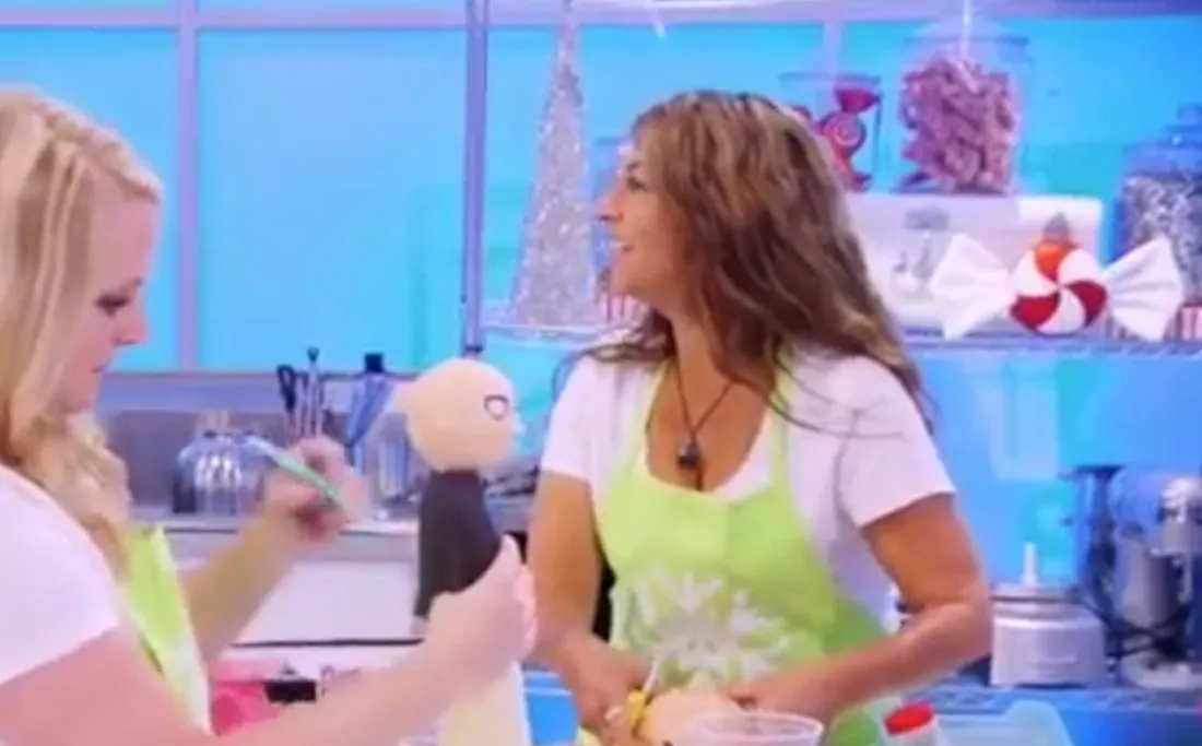 Two women in lime green aprons in the Holiday Wars kitchen, one painting a doll.