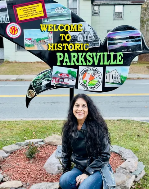 Kim Simons kneeling beside a Parksville welcome sign decorated with local photos and logos.