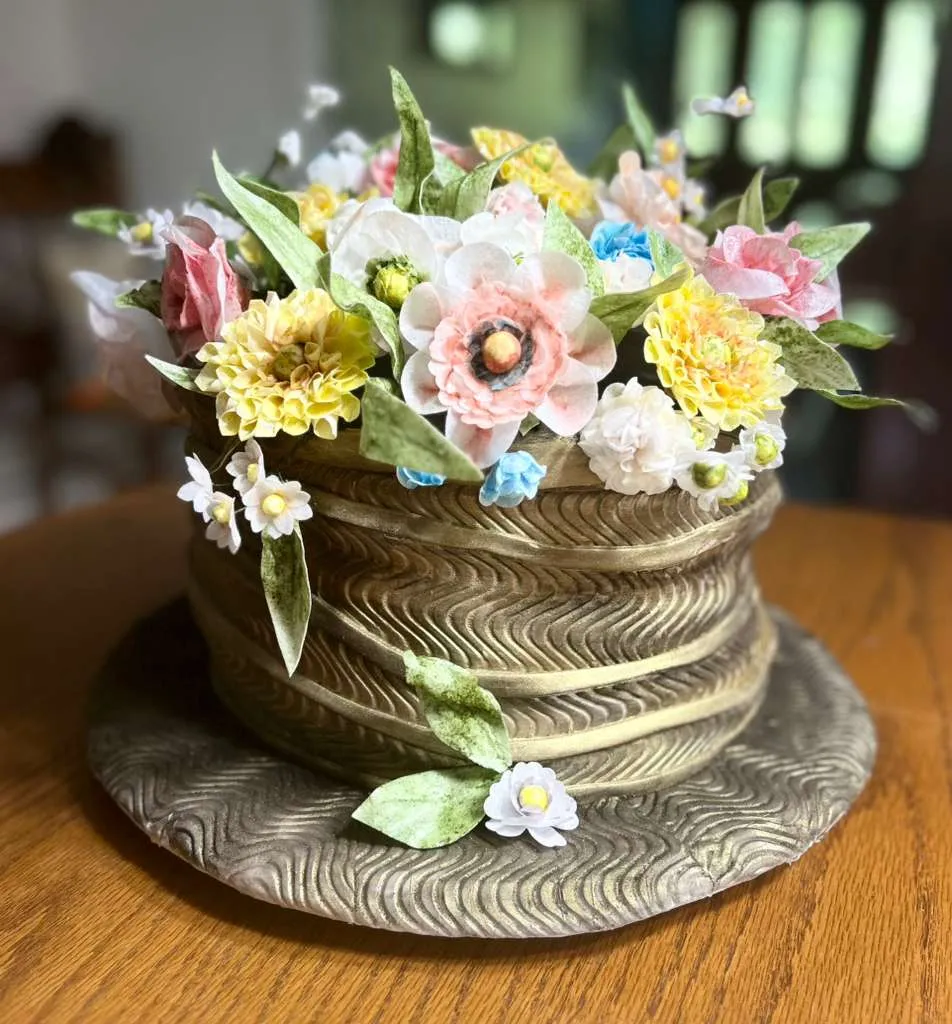 An intricately designed cake styled as a rustic woven basket with metallic gold basket-weave texture, overflowing with handcrafted sugar flowers in soft pastel pinks, yellows, whites, blues, and creams, including detailed roses, daisies, and ranunculus with green leaves and tiny filler blossoms.