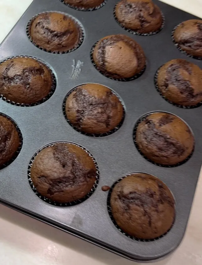 Baked chocolate cupcakes in a dark muffin tin cooling on a light countertop
