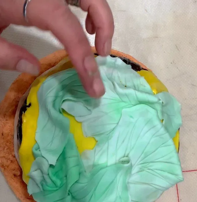 A hand places marbled green and white fondant lettuce onto the burger.