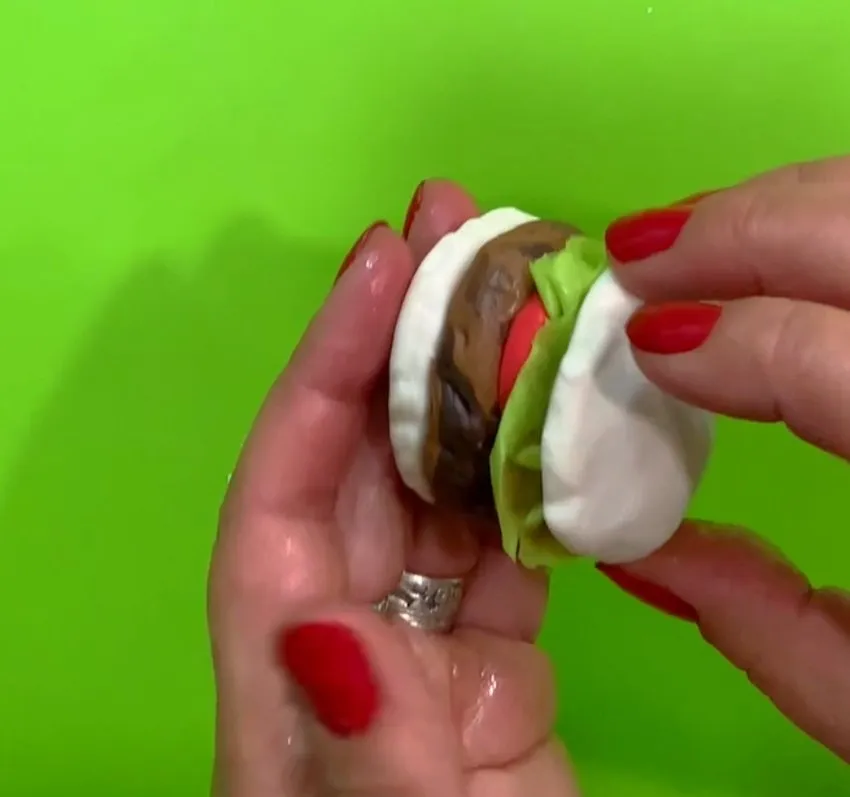 Hands holding a finished fondant hamburger with lettuce and top bun.