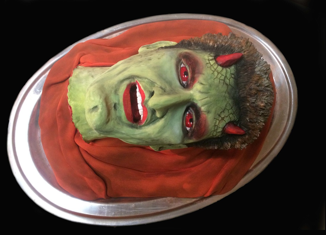 A hyper-realistic cake sculpted to resemble a severed green-skinned demon head with red eyes, small horns, and an open mouth, resting on a red cloth inside a silver platter.