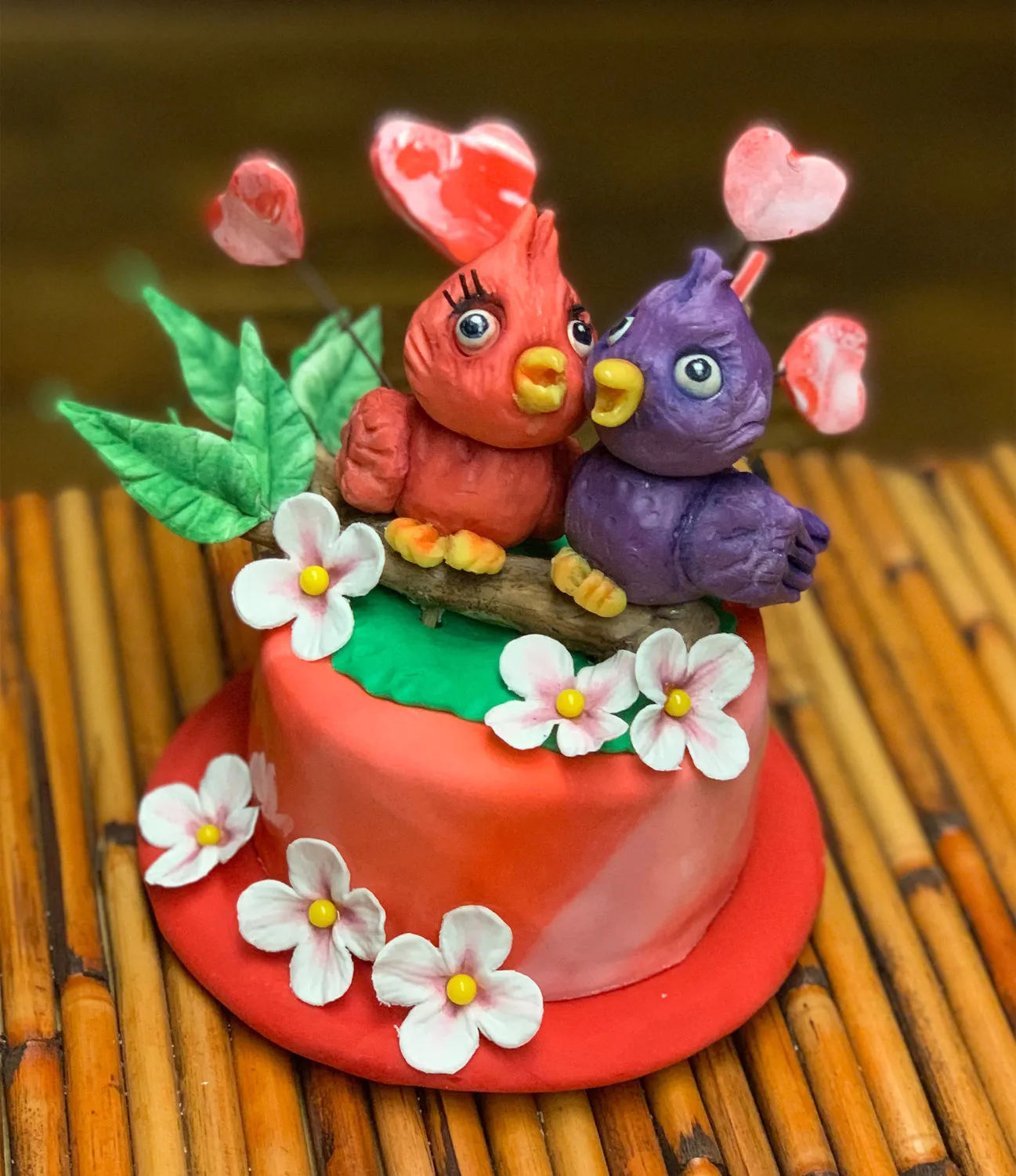 A red fondant cake topped with sculpted lovebird birds on a branch with flowers and hearts