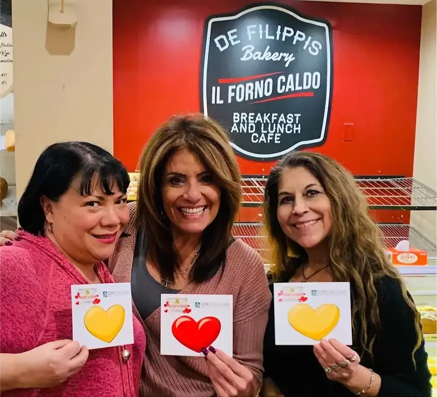 Three women holding heart cards in front of a De Filippis Bakery sign.