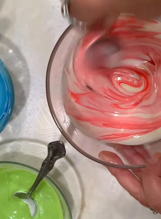 Hand stirring pink, green, and blue melted chocolate in glass bowls.