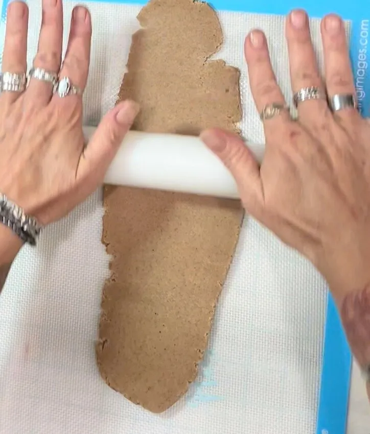 Hands rolling out cookie dough with a rolling pin on a silicone mat.