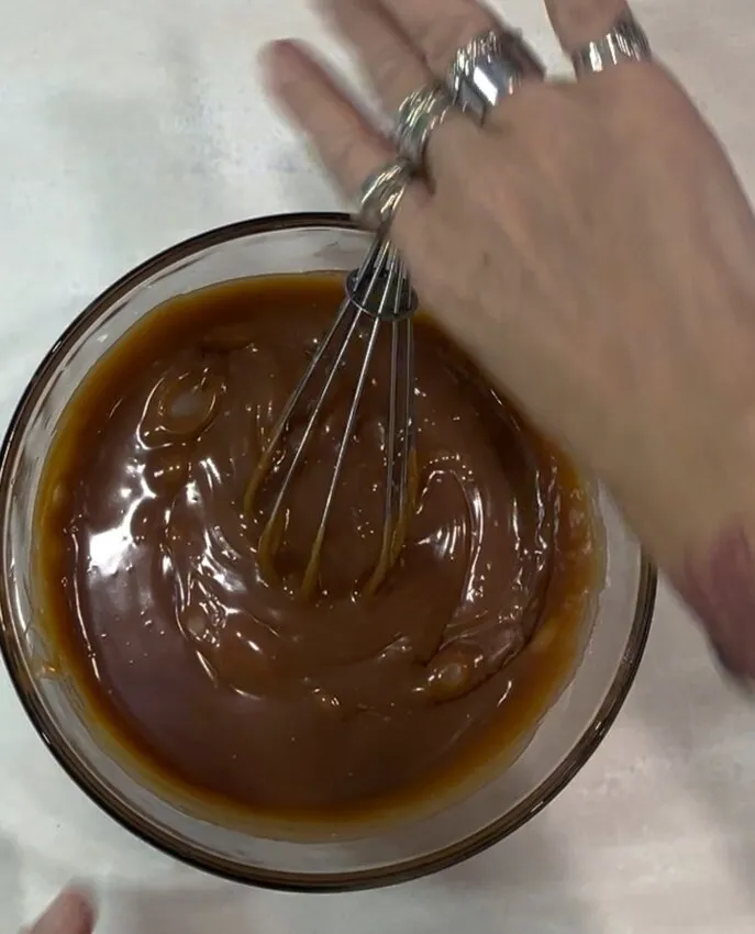 A glass bowl of thick dark chocolate buttercream frosting being mixed with a wire whisk