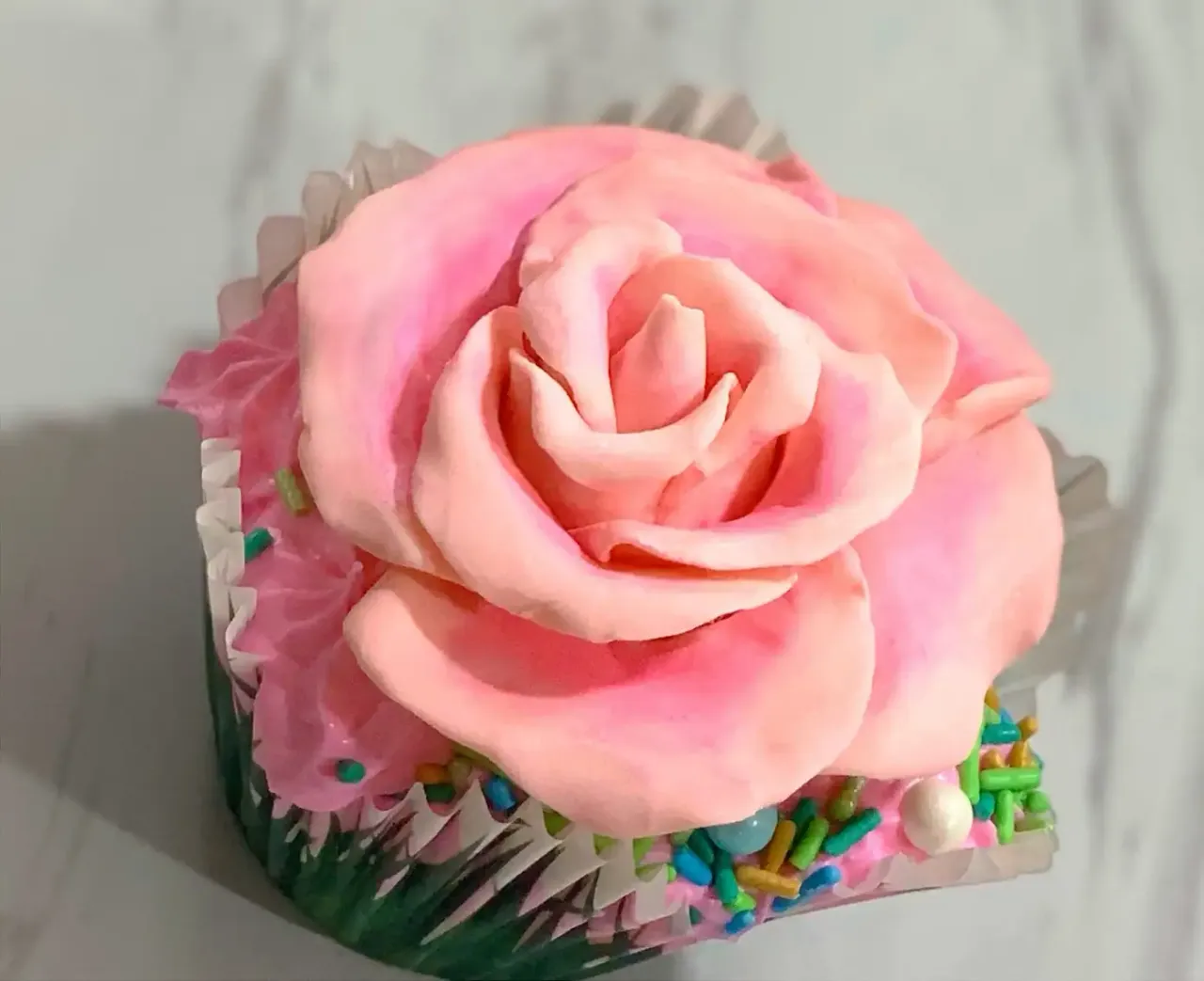 A cupcake topped with a large pink modeling chocolate rose and colorful sprinkles