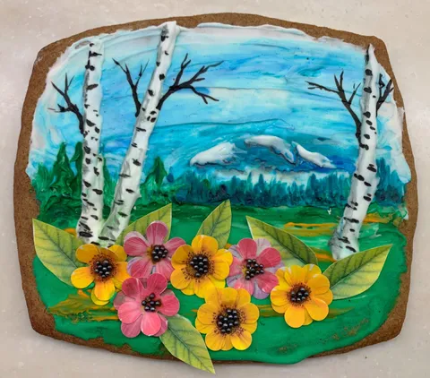 A gingerbread cookie decorated with a landscape painting featuring white birch trees, a blue sky, and pink and yellow flower details