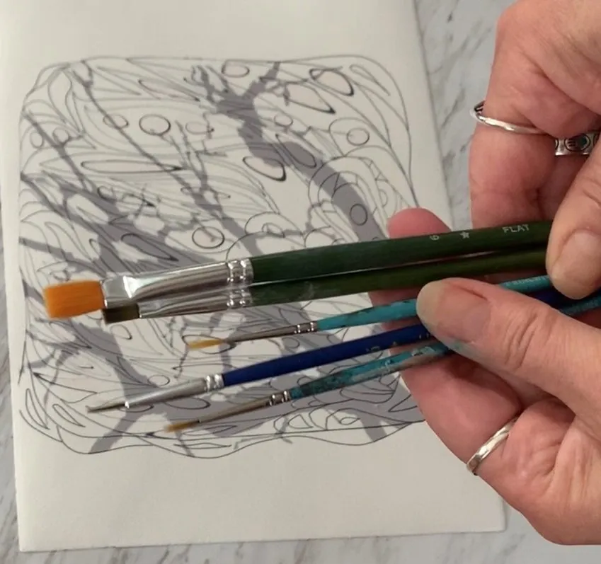 Hand holding paintbrushes over a paper with an intricate black ink line drawing of a butterfly.