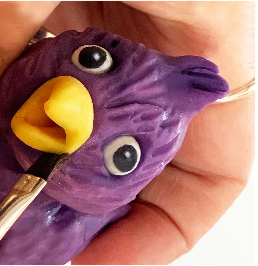 Hand painting details on the purple modeling chocolate lovebird with a brush.