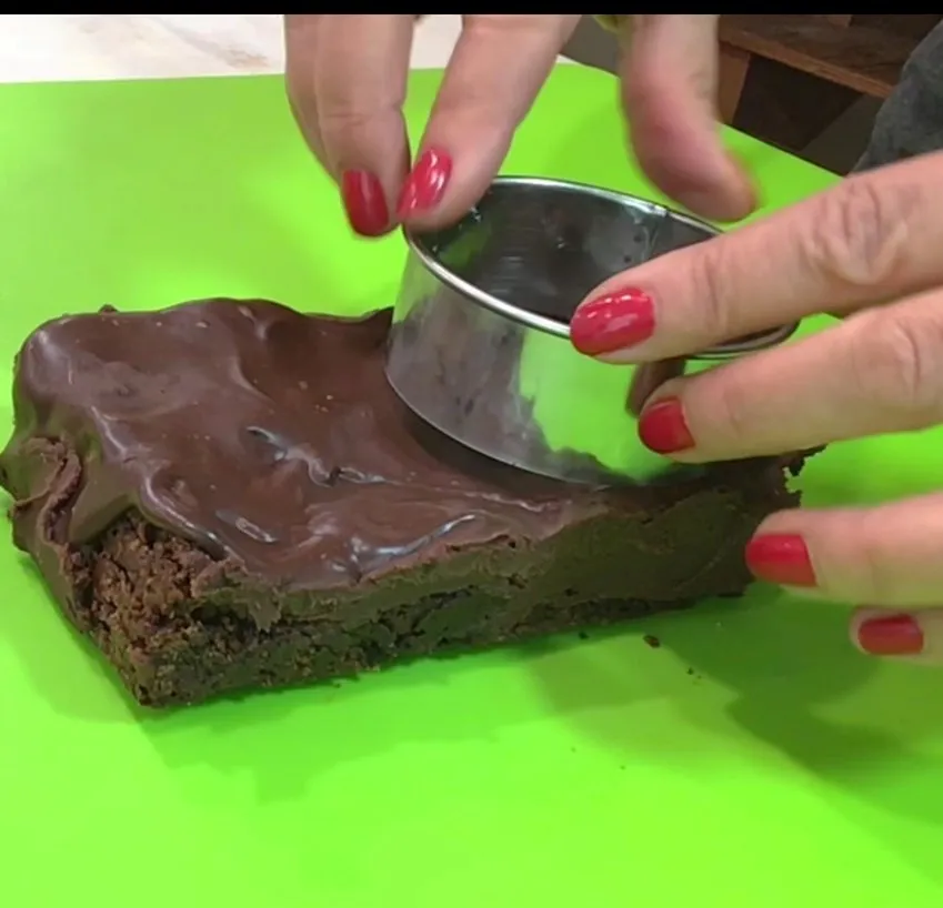 Hands using a round cutter to cut circles from frosted brownies.