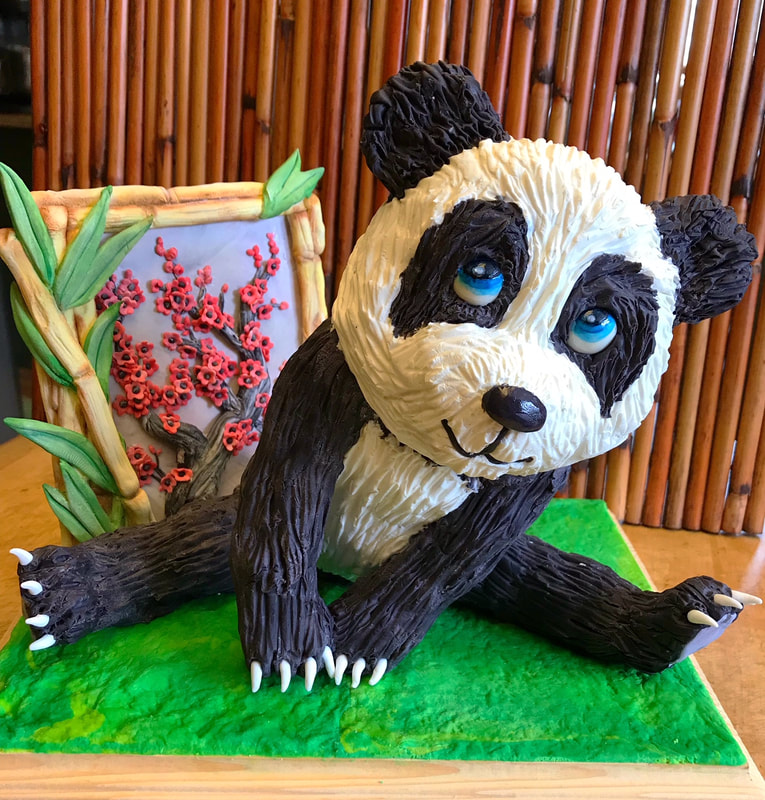 A sculpted panda cake with textured black and white frosting, blue eyes, and white claws sits on a green base next to a decorative bamboo frame featuring red blossoms.