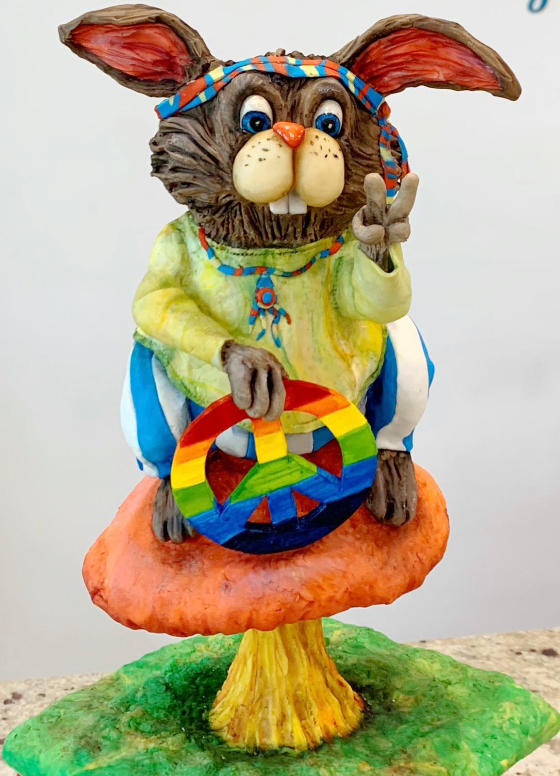 A colorful clay sculpture of a hippie-style rabbit wearing a headband and tie-dye shirt, sitting on a mushroom and holding a rainbow peace sign.