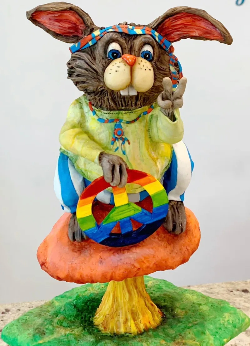 A colorful clay sculpture of a hippie-style rabbit wearing a headband and tie-dye shirt, sitting on a mushroom and holding a rainbow peace sign.