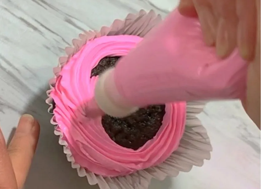 Hand piping pink buttercream swirl onto a chocolate cupcake.
