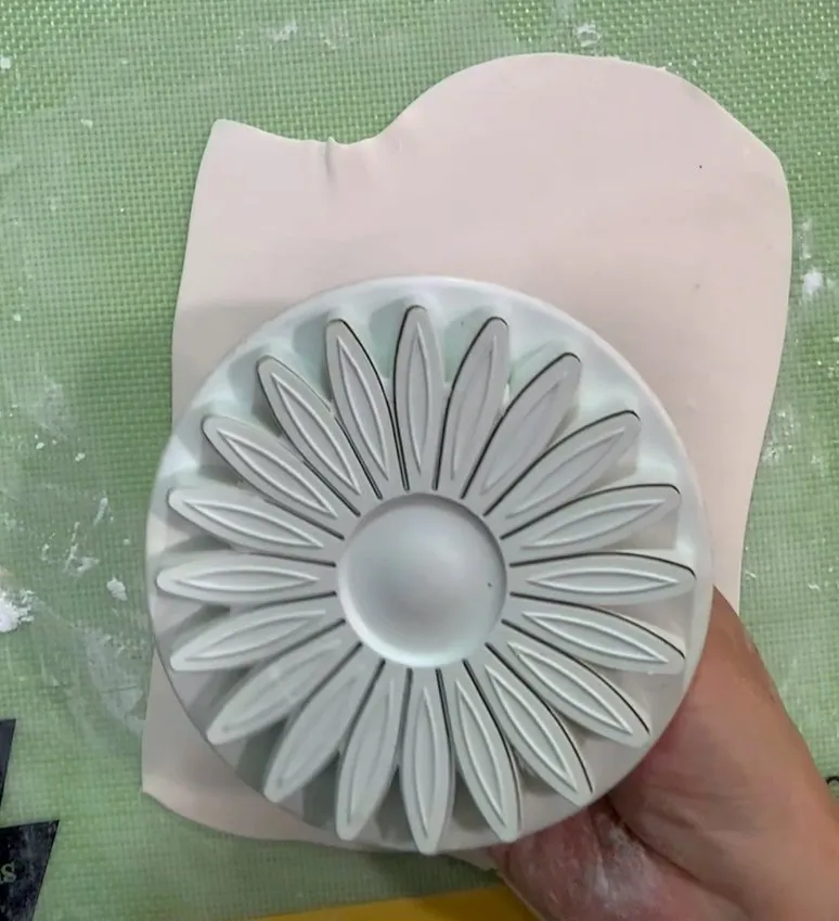 Hand pressing a sunflower cookie cutter into rolled gumpaste on a mat.