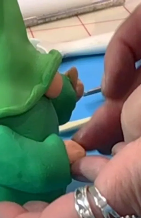 A hand uses a cake tool to shape fondant arms on the gnome body.