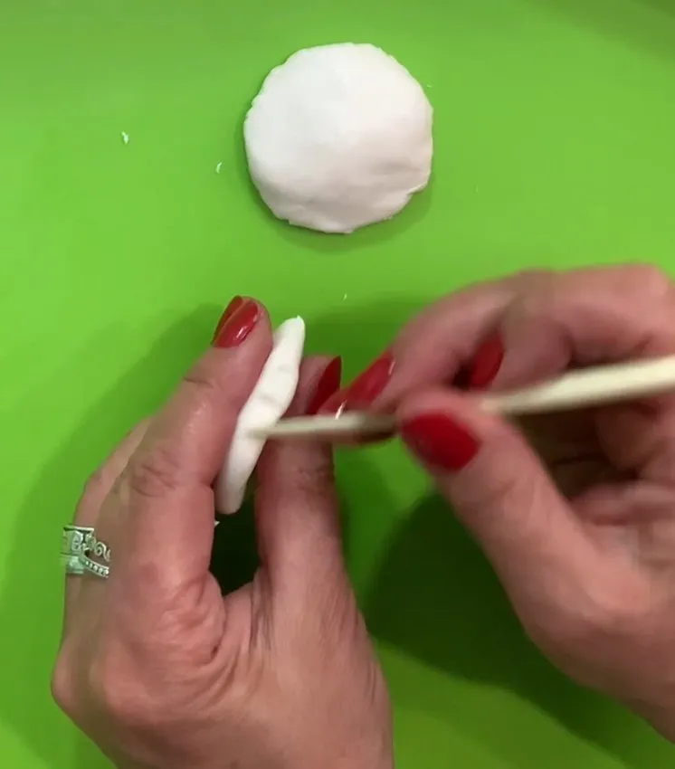 Hands shaping white fondant into a hamburger bun on a green mat.