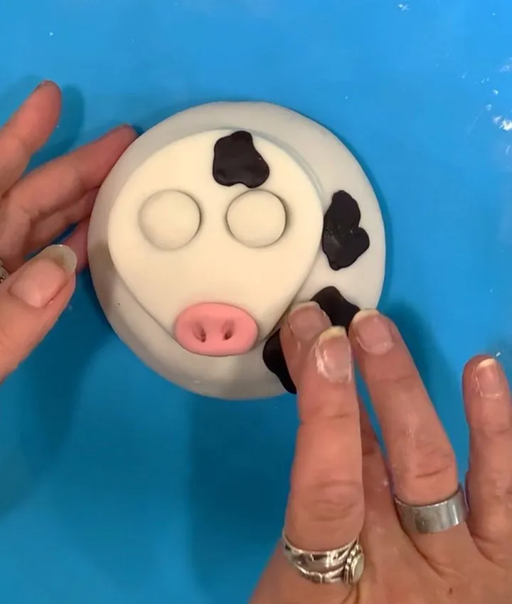 Decorating a fondant cow face with black spots, eyes, and a pink snout.