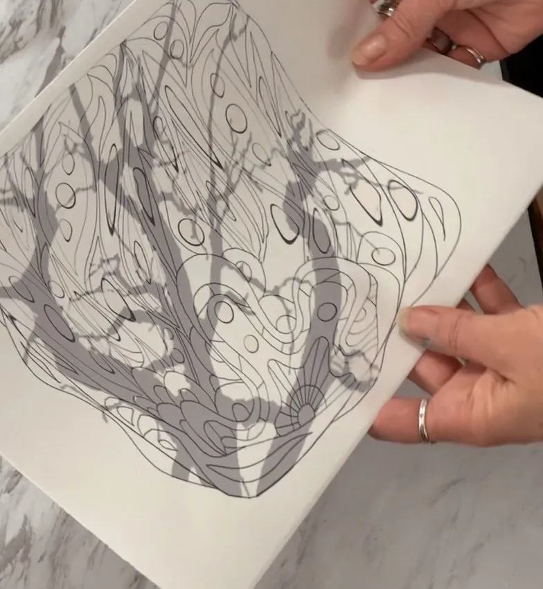 Hands holding a paper with an ink line drawing of a stylized tree with swirling branches.
