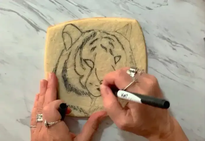 Hand drawing a tiger face sketch on a sugar cookie with a marker.
