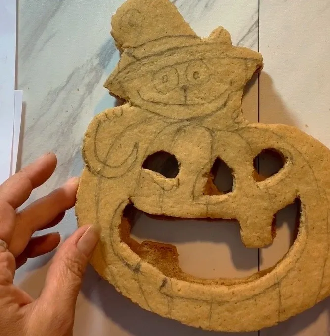 A hand holds a Halloween cookie with a sketched cat-on-pumpkin design.