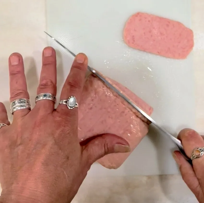 Hands slicing a block of canned meat on a cutting board.