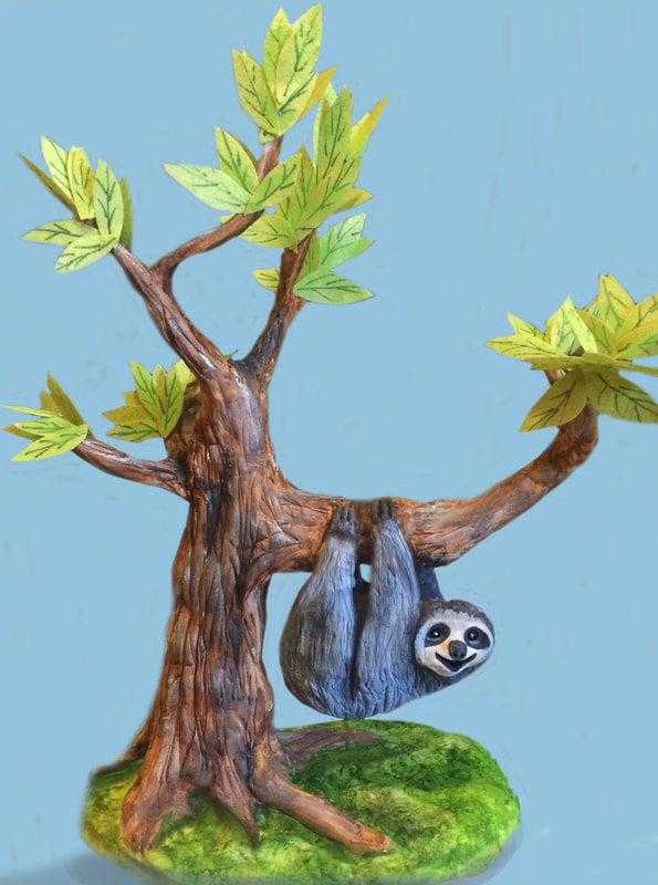A painted figurine of a smiling sloth hanging upside down from a brown tree branch with green leaves, set on a mossy green base against a light blue background.