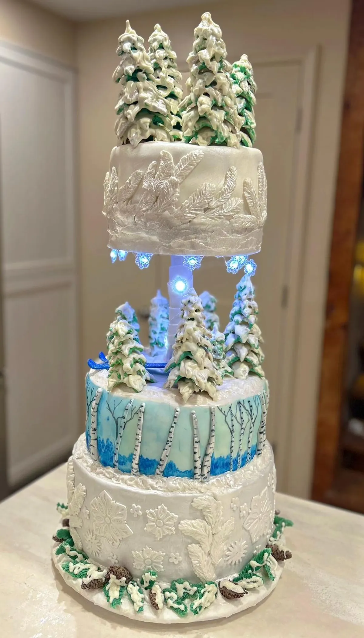 A three-tiered winter-themed cake features white fondant decorated with embossed snowflakes and pine branches, a middle tier painted with a blue birch forest scene, and piped sugar pine trees dusted with white "snow" on the top and bottom tiers, illuminated by blue LED lights underneath the upper layer.