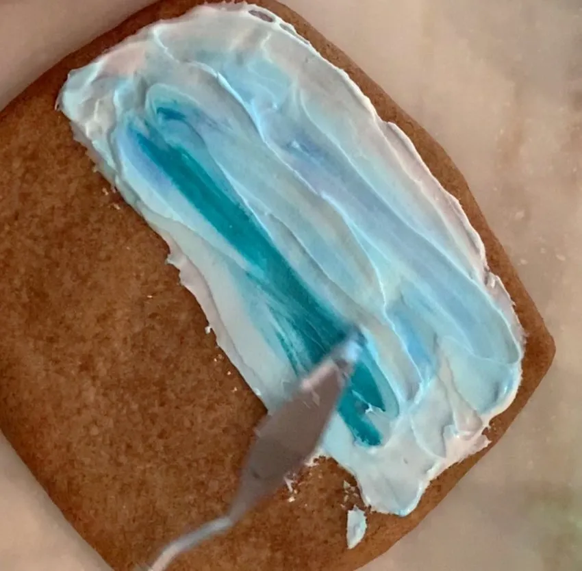 Knife spreading blue and white royal icing sky onto a square cookie.