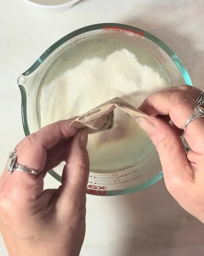 Hands sprinkle gelatin powder over water in a glass measuring cup.