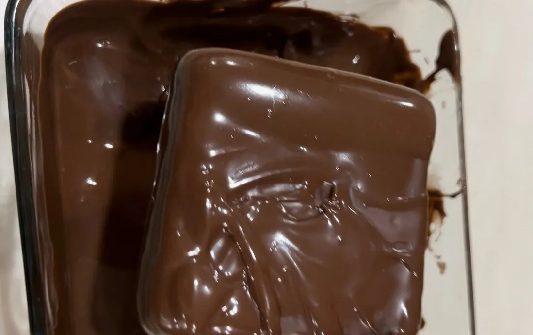 Avocado-banana frozen treats being coated with melted dark chocolate in a glass bowl