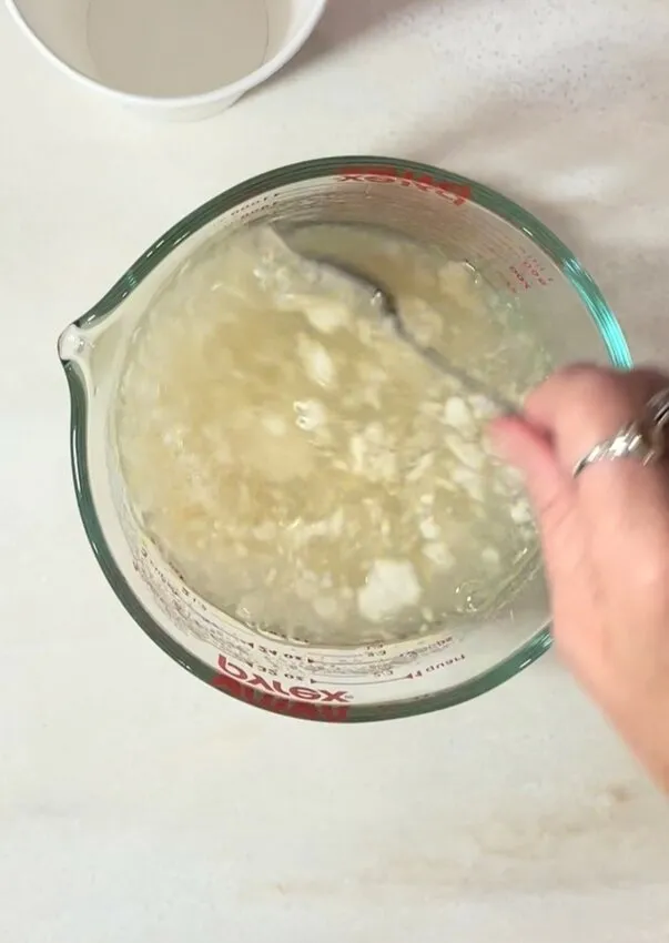 A hand stirs the gelatin mixture as it begins to set in a glass measuring cup.