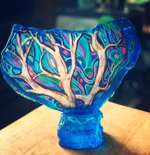A translucent blue sugar sculpture featuring a painted tree with intricate, colorful swirling patterns in the background, mounted on a matching blue sugar base.