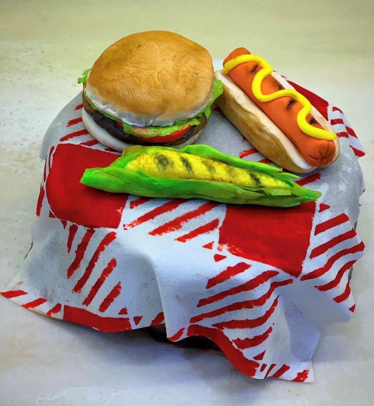 A cake decorated as a picnic scene with fondant hamburger, hot dog, and corn on a checkered tablecloth