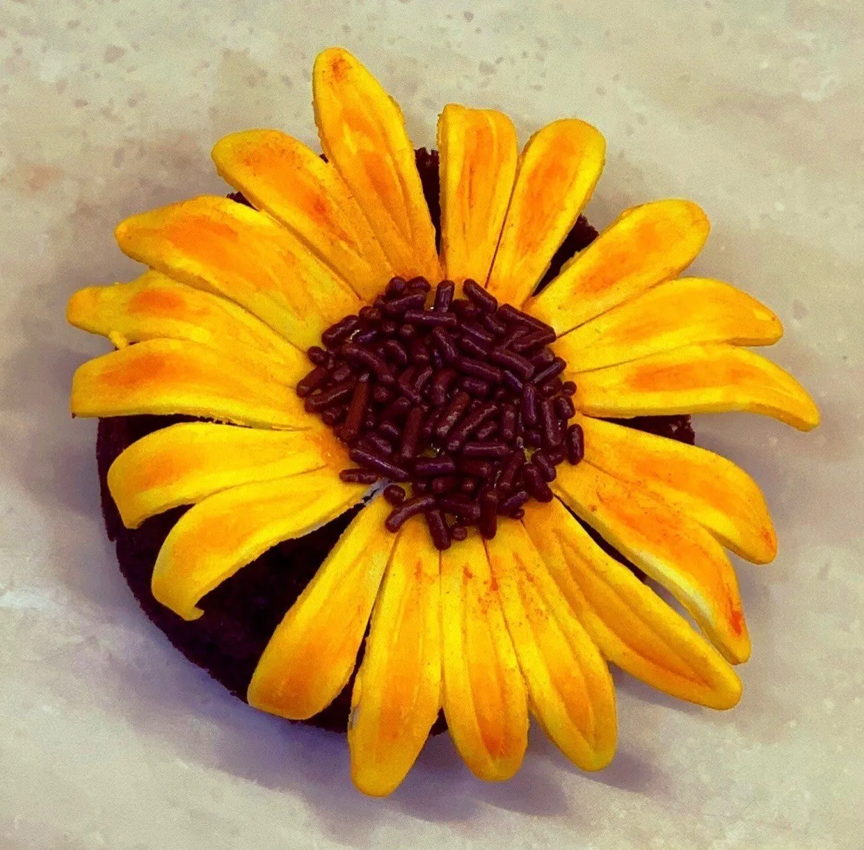 A brownie topped with yellow frosting piped into a sunflower shape with dark sprinkles in the center