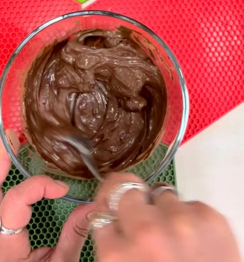 Hand stirring melted chocolate in a glass bowl on a silicone mat.