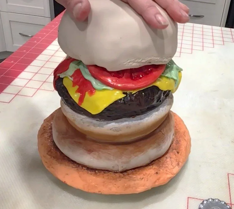 A hand places the fondant-covered top bun onto the burger cake.