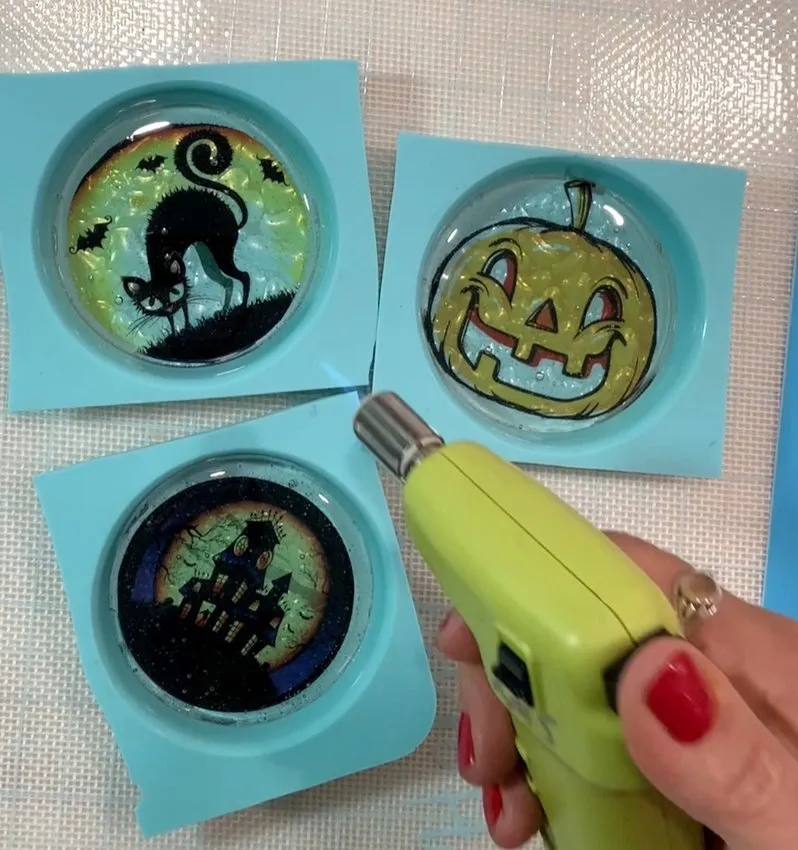 A hand uses a blow torch to remove bubbles from isomalt lollipops in molds.