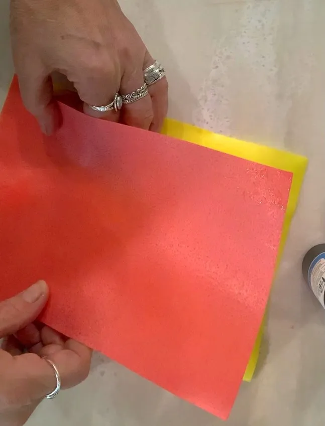 Hands holding a sheet of orange wafer paper over a yellow sheet.