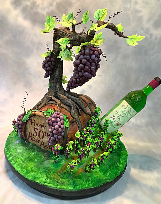 An elaborate sculpted cake features a grapevine with purple clusters growing from a wooden barrel labeled "Happy 50th Rose Ann," accompanied by a green wine bottle and a base resembling a grassy vineyard.