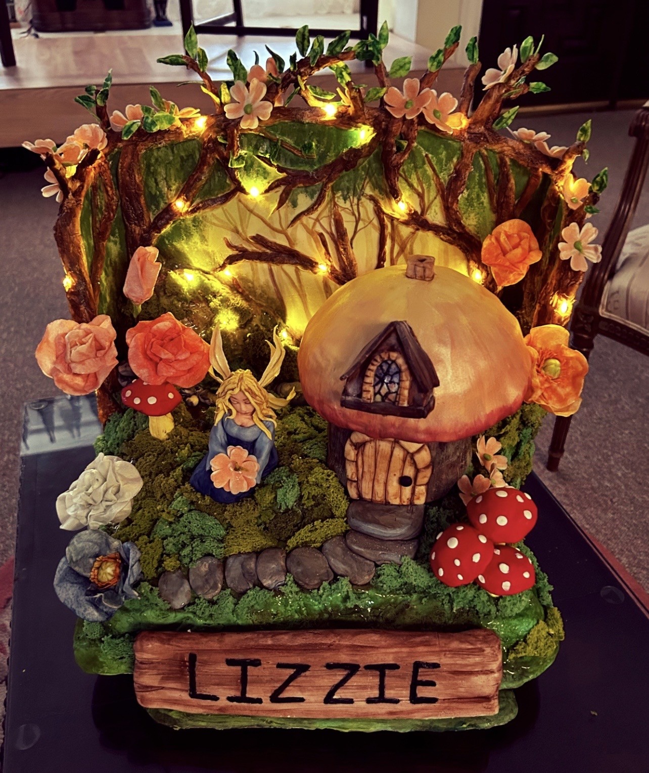 A whimsical cake designed as a magical forest scene centered around a mushroom house with an orange cap, wooden door and window, nestled among lush green moss-like frosting, vibrant red-and-white spotted mushrooms, sugar flowers in peach, coral, and cream, a fairy figure with golden wings, and a rustic wooden sign bearing the name "LIZZIE."